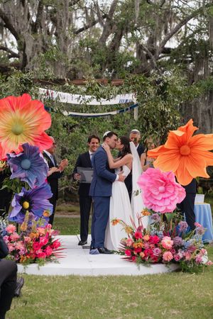 Newlyweds kiss at altar surrounded by giant colorful paper flowers at outdoor garden wedding.