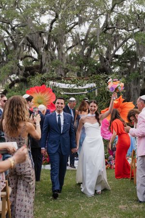 Joyful newlyweds exit outdoor wedding ceremony under floral arch as guests celebrate with colorful decor.