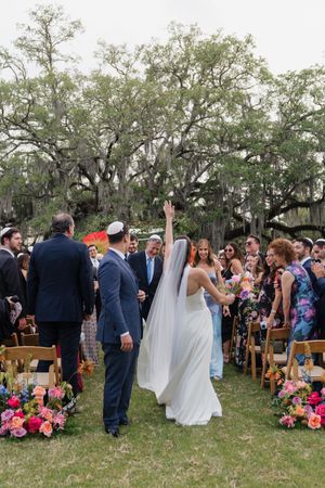 Bride in white gown and veil raises hand joyfully with groom in navy suit after outdoor ceremony under oak trees.