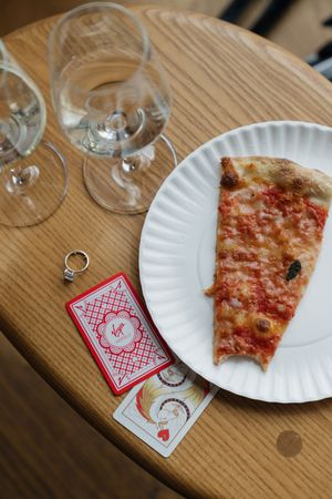 Wedding ring beside playing cards, pizza slice on paper plate, and empty wine glasses on a wooden table.
