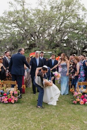 Newlyweds sharing romantic dip kiss while walking down aisle at outdoor Jewish wedding ceremony.