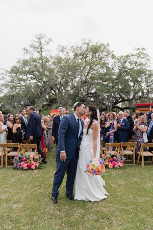 Newlyweds share first kiss walking back down the aisle at vibrant outdoor wedding with colorful floral arrangements.
