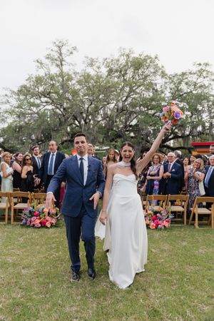 Joyful bride raises bouquet while walking with groom down aisle after outdoor wedding ceremony with guests celebrating.