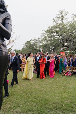 Wedding guests in colorful formal attire walking across lawn at outdoor ceremony celebration.