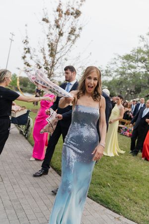 Woman in silver sequin gown smiles at outdoor wedding celebration with colorful-dressed guests.