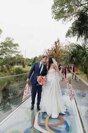 Newlyweds share a kiss on a glass bridge with colorful mural at outdoor wedding venue.