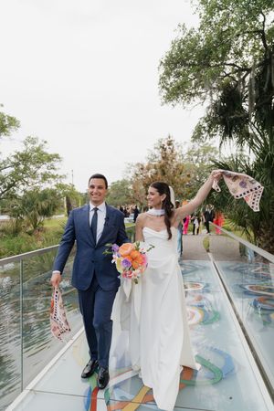 Joyful newlyweds walk across colorful glass bridge waving scarves after outdoor wedding ceremony.
