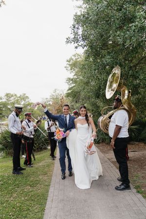 Bride and groom walk joyfully with a brass band during their wedding celebration outdoors.