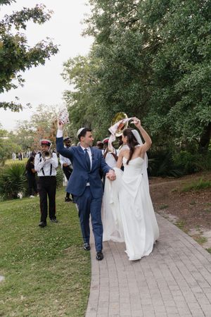 Bride and groom celebrate with second line parade on garden path surrounded by lush greenery after wedding ceremony.