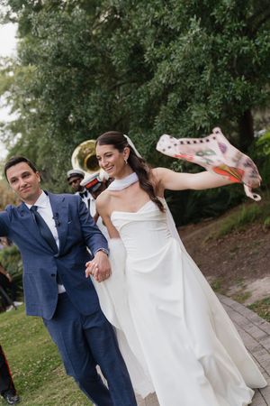 Smiling bride and groom exit ceremony as bride waves decorated sneaker at outdoor wedding.