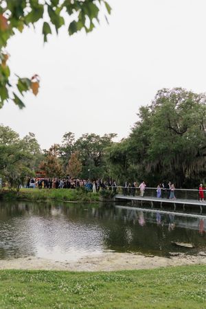 Wedding guests cross scenic bridge over tranquil pond surrounded by moss-draped trees at outdoor venue.