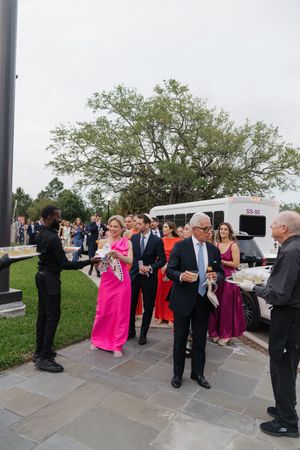 Wedding guests in formal attire including pink and red gowns arrive via shuttle bus at outdoor venue.