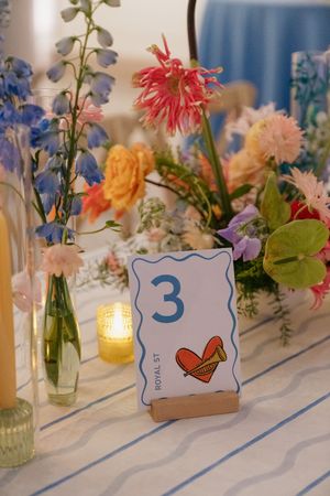 Wedding table number 3 card with heart illustration surrounded by colorful wildflower arrangements and candles.
