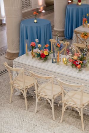 Elegant wedding reception table with colorful floral centerpieces, blue linens, and wooden cross-back chairs.