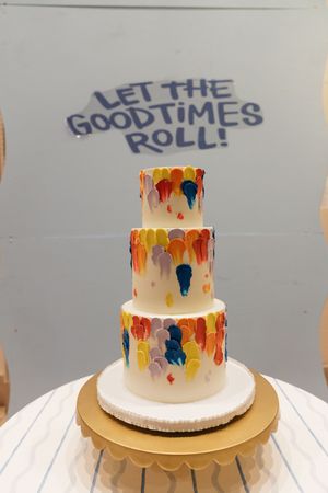 Three-tier white wedding cake with colorful brushstroke details beside Let The Goodtimes Roll sign.