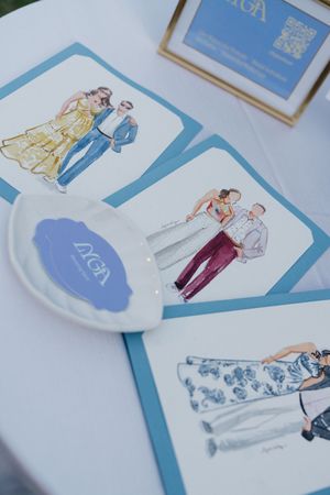 Watercolor fashion illustrations displayed on blue-bordered cards beside a branded plate at an elegant event table.