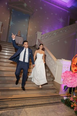 Bride and groom descend grand staircase at wedding reception, groom pointing excitedly in navy suit, bride in white gown.