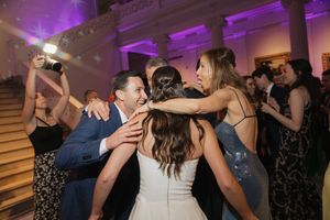 Bride and groom share a joyful group hug with guests on a crowded dance floor under purple wedding reception lighting.
