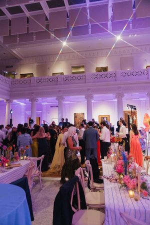 Guests mingle in a grand ballroom with purple lighting, ornate columns, and colorful floral centerpieces.