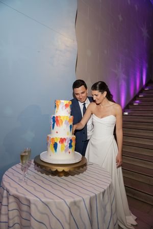 Bride and groom cut colorful painted three-tier wedding cake at elegant reception venue.