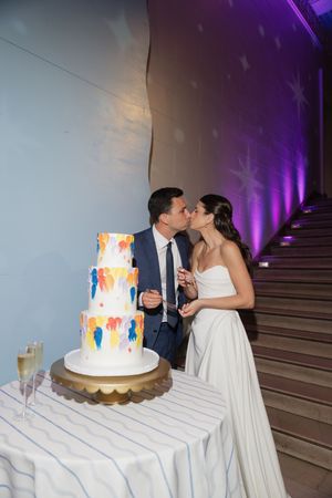 Bride and groom kiss beside a three-tier white wedding cake with colorful painted details near a staircase.