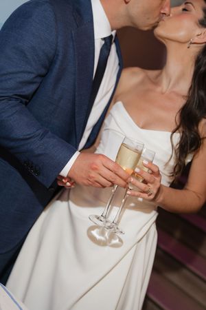 Bride and groom share a kiss while holding champagne flutes together during their wedding reception toast.