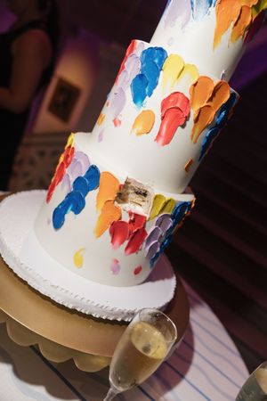 Two-tier white wedding cake decorated with colorful painted brushstroke designs beside champagne flute.