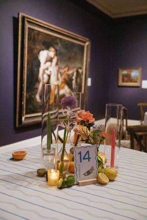 Wedding table centerpiece with colorful flowers, candles, citrus fruits, and a number 14 card in an art museum.