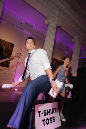 Groom in navy suit participates in a fun t-shirt toss activity at a lively wedding reception with purple lighting.