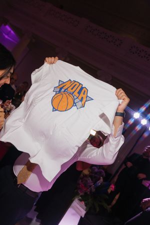 Person holds up a white NOLA basketball-themed t-shirt at an indoor celebration event.