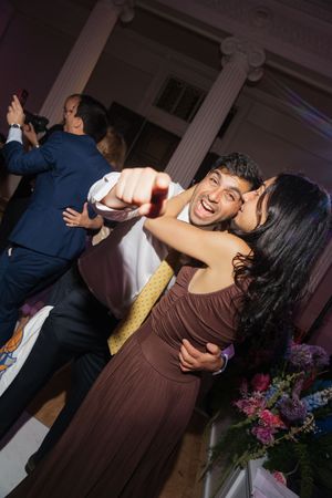 Joyful couple dancing and pointing at camera during a festive indoor wedding reception.