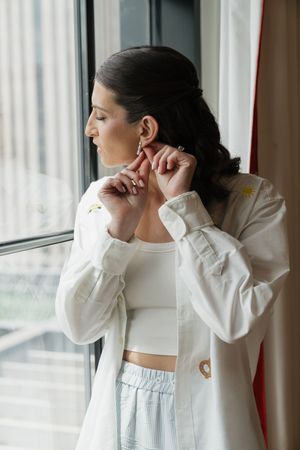Bride putting on earrings by window in white jacket and crop top while getting ready on wedding day.