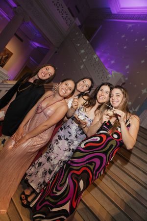 Five women in elegant dresses smile and pose together at a festive wedding reception with purple ambient lighting.