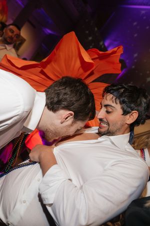 Two men in white shirts laugh and dance energetically under orange fabric at wedding reception.