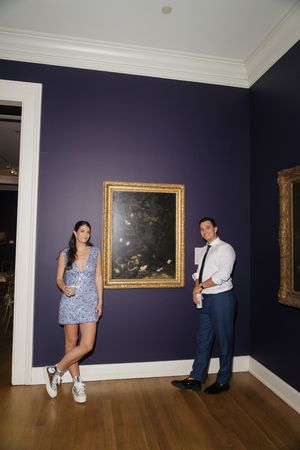 Couple posing beside gold-framed painting on deep purple wall in art gallery venue.