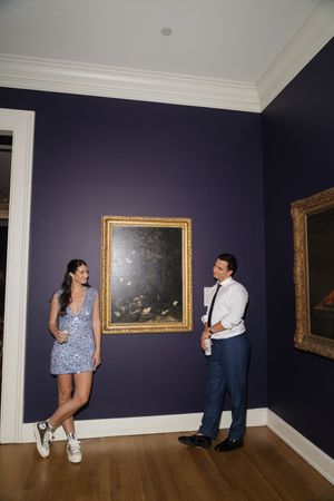 Couple in casual attire laughing together beside a gold-framed painting on a purple gallery wall.