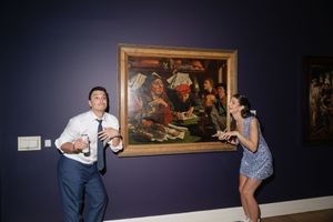 Couple posing playfully beside a large classical painting on a dark blue wall in an art museum.