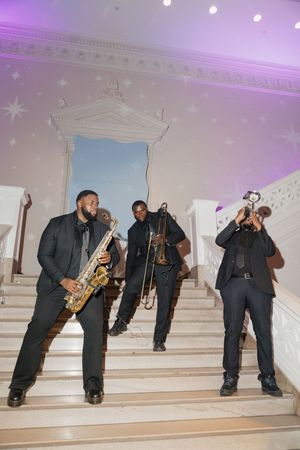 Three musicians in black suits perform on grand staircase with saxophone, trombone, and trumpet at wedding.