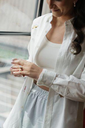 Bride smiling while adjusting her gold ring, wearing a white open shirt over a crop top and blue striped skirt.