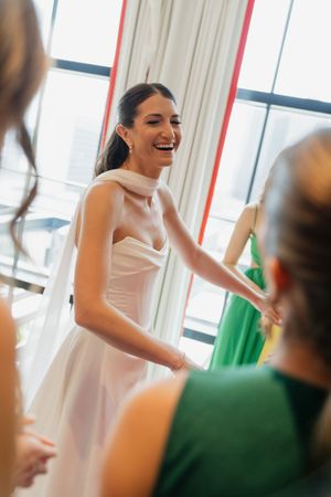 Bride laughing in white wedding gown during dress fitting with bridesmaids in green dresses.