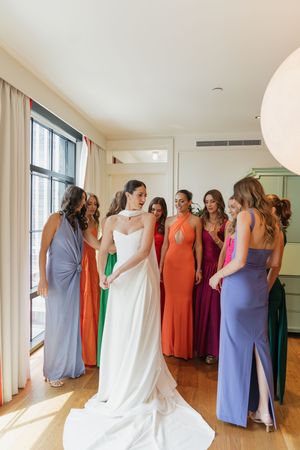Bride in white gown surrounded by bridesmaids in colorful dresses during wedding preparations in a bright room.