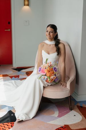 Bride in white gown holds vibrant bouquet while seated in elegant chair.