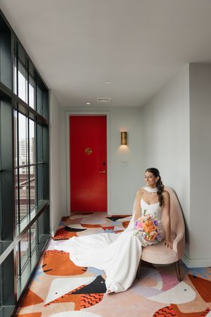 Elegant bride in white gown sitting on blush chair in modern hotel room with red door and bold carpet.