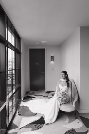 Black and white portrait of bride in wedding gown seated by large windows holding bouquet.