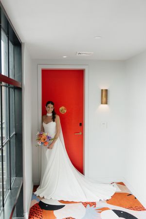 Bride in elegant white gown with long train poses in front of vibrant red door with modern gold sconce.
