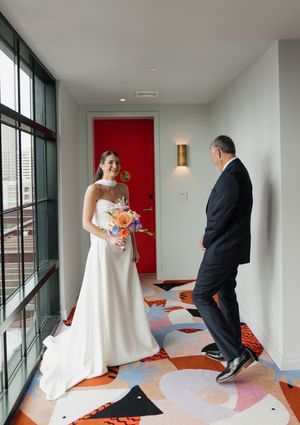 Bride in strapless white gown holding colorful bouquet meeting her father in a vibrant patterned hallway.