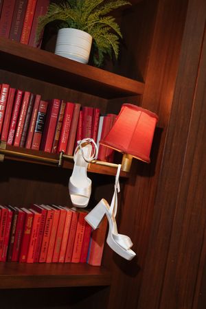 White bridal heels hanging on a lamp beside red books on a wooden bookshelf with a green plant.