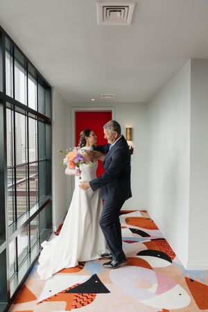 Bride in elegant white gown receiving a warm embrace from her father in a decorated hotel hallway.