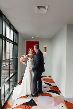 Bride in white gown sharing first look moment with father in hotel corridor with colorful carpet.