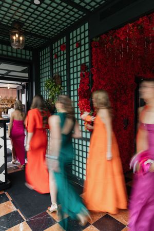 Blurred motion of women in colorful formal gowns walking past a red floral wall at an elegant event.